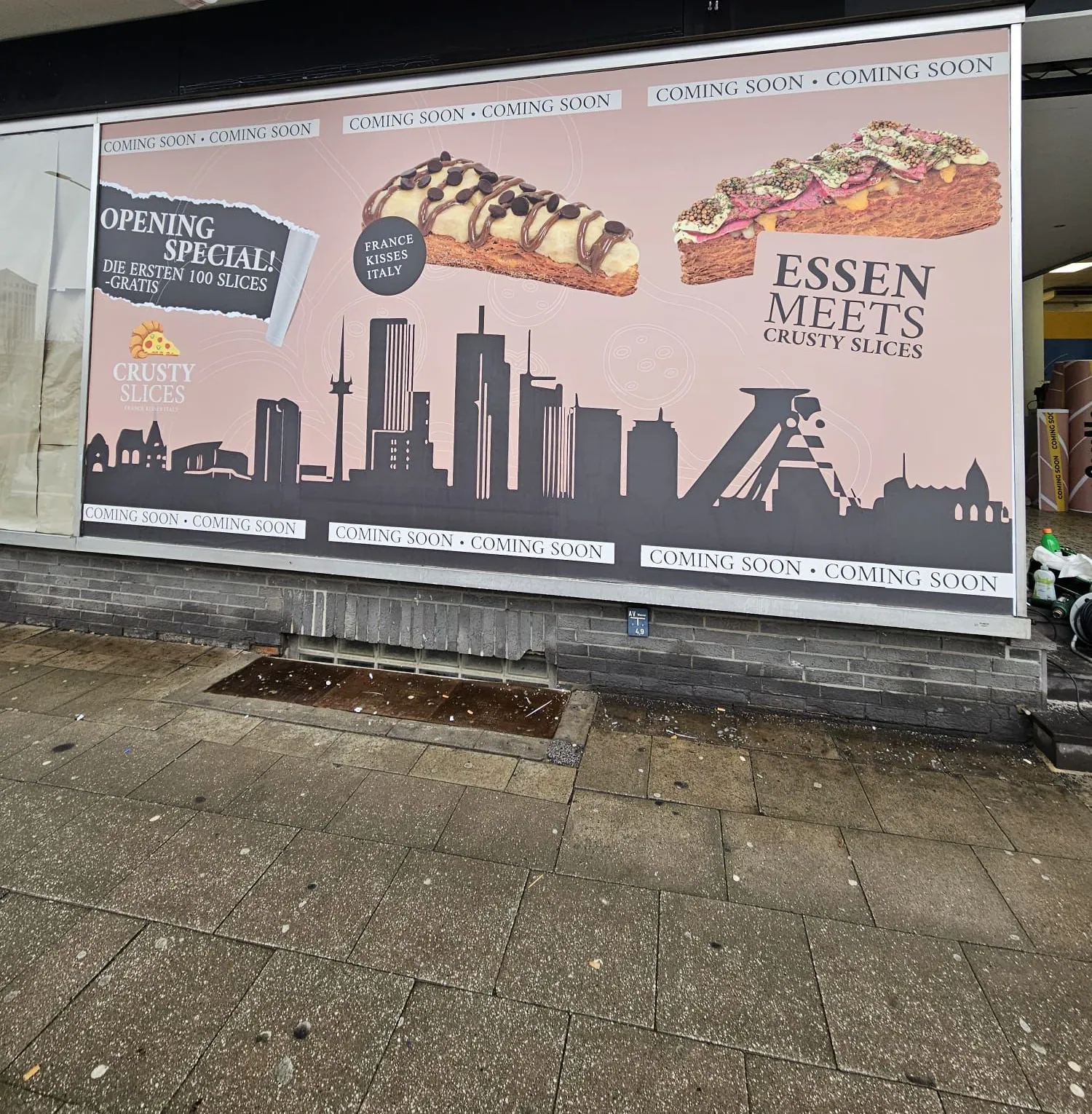 Großflächig beklebtes Schaufenster in Essen mit Stadtsilhouette, zwei Croissant-Pizza-Stücken und dem Hinweis Coming soon
