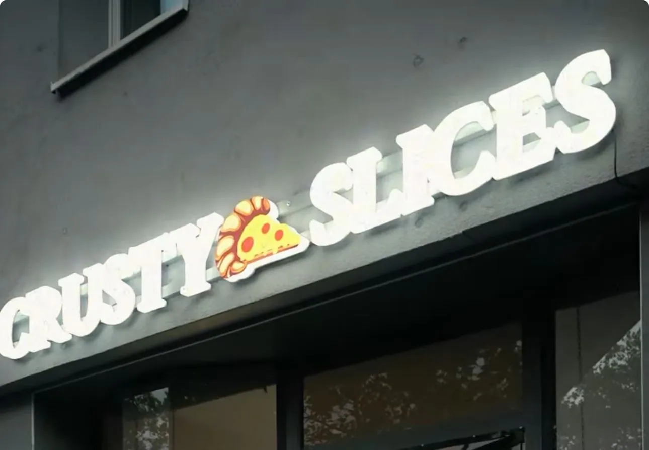 Weiß leuchtender Schriftzug Crusty Slices mit Pizza-Symbol an einer grauen Ladenfassade über dem Eingang