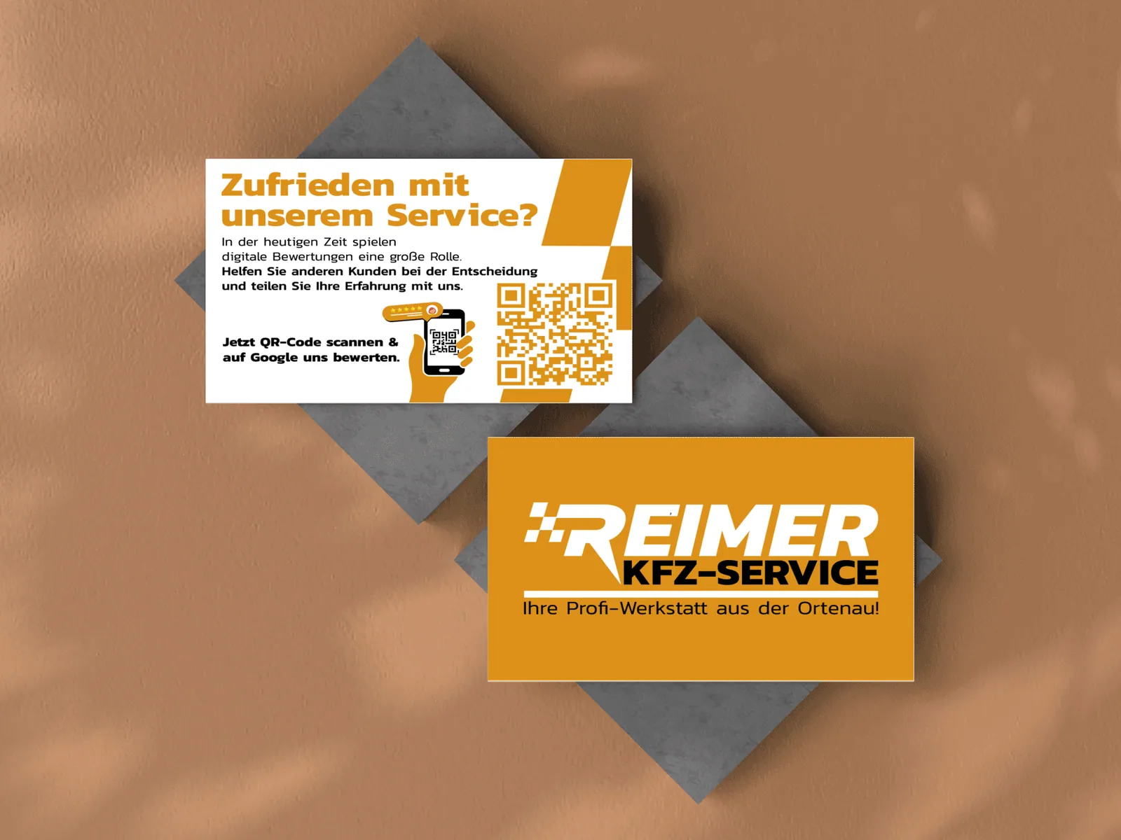 Orange Visitenkarte mit Logo von KFZ-Service Reimer und weiße Google-Bewertungskarte mit QR-Code auf grauen Platten vor einer Steinwand